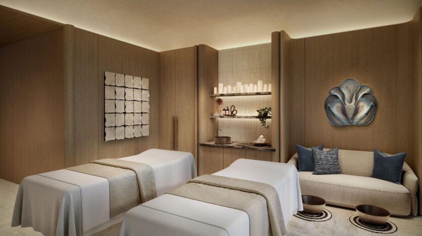 The serene spa room at Four Seasons Private Residences features two massage tables, beige linens, a modern sofa with blue pillows, a shelf displaying candles and decorative items, and soft, warm lighting to create a truly relaxing atmosphere.
