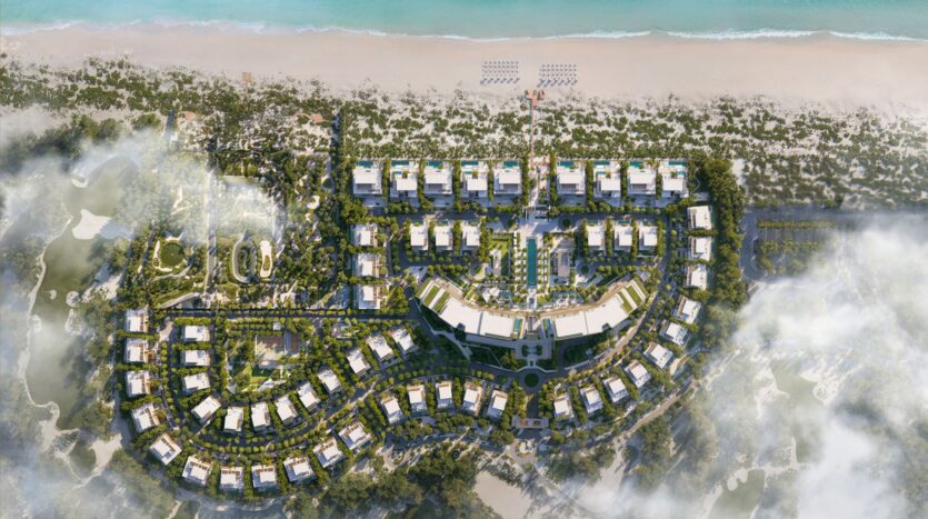 An aerial view of the luxurious Four Seasons Private Residences beachfront resort, consisting of multiple white buildings arranged in curved and straight rows, surrounded by lush greenery, walking paths, and a sandy beach bordering the turquoise waters of the ocean.