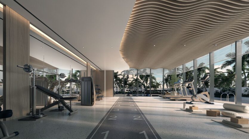 La salle de sport moderne des résidences Meriden Beach est dotée de grandes fenêtres donnant sur les palmiers. Elle comprend des machines d'exercice, des poids libres, des tapis de course, des tapis de yoga et une piste d'athlétisme. Le plafond ondulé et la lumière naturelle créent une atmosphère accueillante.