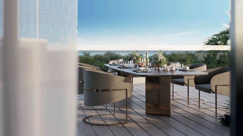 Salle à manger moderne en plein air sur une terrasse en bois des résidences Meriden Beach, avec une table rectangulaire et des chaises grises élégantes, donnant sur une végétation luxuriante et un ciel bleu limpide.