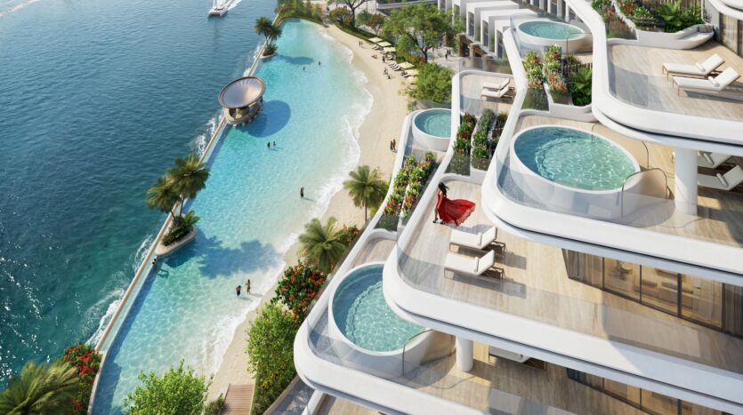 Aerial view of River Cove Residences, a luxury waterfront complex featuring private pools on the balconies, a sandy beach, palm trees, and people swimming in a blue lagoon by the sea. A yacht is visible in the water.