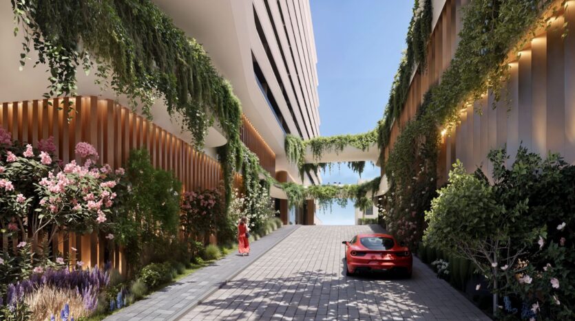 A modern courtyard at River Cove Residences, featuring large buildings adorned with lush greenery and flowers. A red sports car is parked on the stone path, while a person dressed in red walks toward the open blue sky beyond the courtyard.