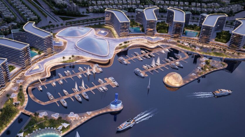 An aerial view of a modern marina featuring luxury yachts, the curved, futuristic buildings of River Cove Residences, and illuminated walkways. Several boats are moored, and a few are sailing near unique architectural structures.