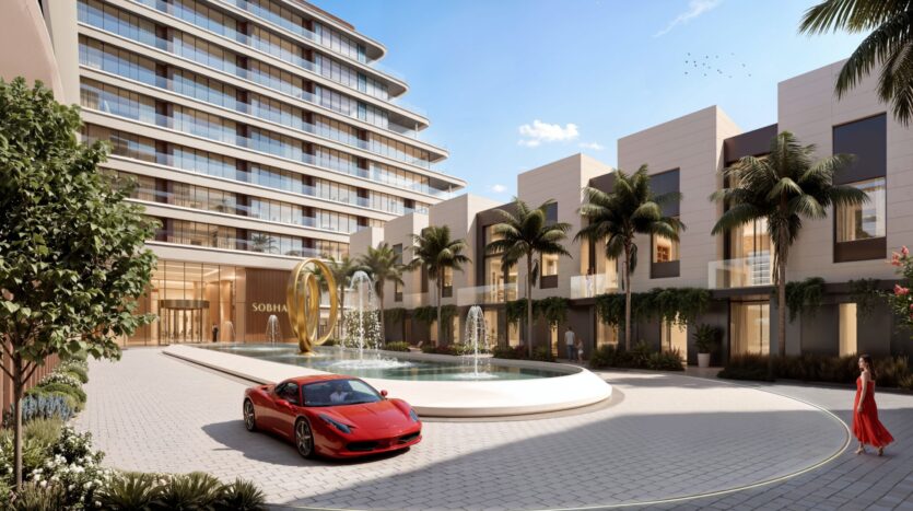 The entrance to a modern luxury hotel at River Cove Residences features a water fountain, palm trees, a red sports car parked out front, and a woman in a red dress walking nearby under a clear blue sky.