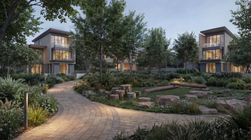 Ein Landschaftsgarten mit einem gewundenen Steinweg, umgeben von Bäumen, Sträuchern und modernen zweistöckigen Gebäuden in der Abenddämmerung im „The Orchard by Sobha“. Holzscheite und Steine schaffen in dieser friedlichen Umgebung einen gemütlichen Platz zum Verweilen.