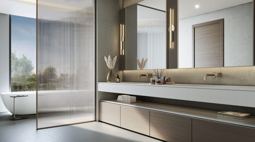 Modernes Badezimmer im „The Orchard by Sobha“ mit einem Doppelwaschtisch, großen Spiegeln, warmer Beleuchtung, Einbauschränken und einer freistehenden Badewanne neben einem großen Fenster. Die neutralen Farbtöne und das minimalistische Design schaffen eine klare und ruhige Atmosphäre.