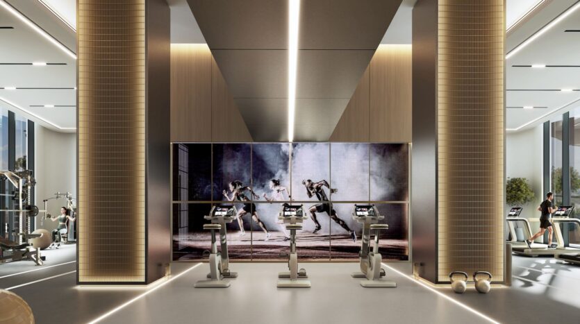 Moderner Fitnessraum im „The Orchard by Sobha“ mit hohen Säulen, Heimtrainern vor einer großen Leinwand, auf der Boxkämpfe gezeigt werden, und verschiedenen Fitnessgeräten an den Seiten. Der Raum ist hell, geräumig und gut beleuchtet und besticht durch ein zeitgemäßes Design.