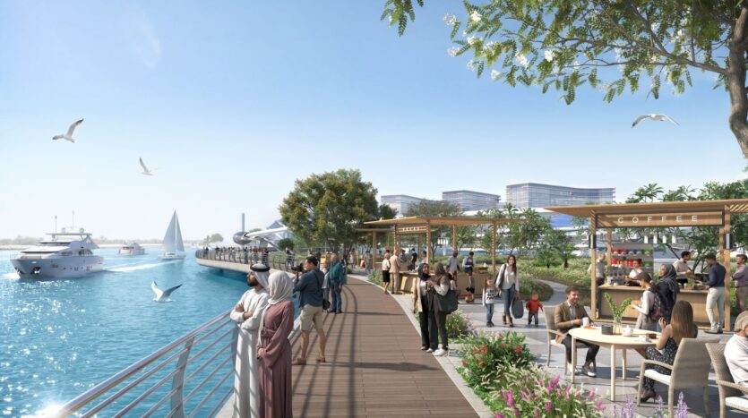 People stroll along The Terraces by Sobha, a modern waterfront promenade lined with cafés, trees, and outdoor seating. Boats glide across the blue water while seagulls take flight, creating a lively scene under a clear, sunny sky.