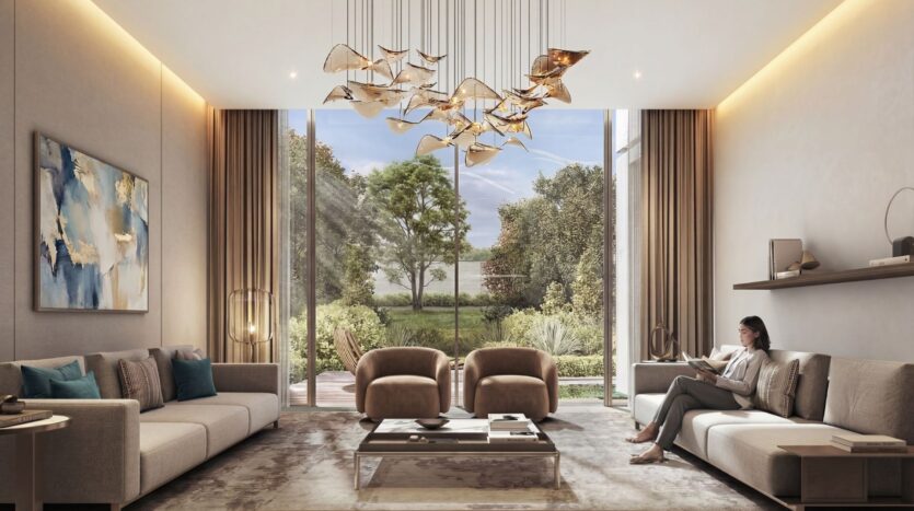 A modern living room decorated in neutral tones at The Terraces by Sobha features large windows overlooking a garden. Elegant furniture, abstract artwork, and a gold chandelier adorn the space. A woman is sitting on a sofa reading a book.