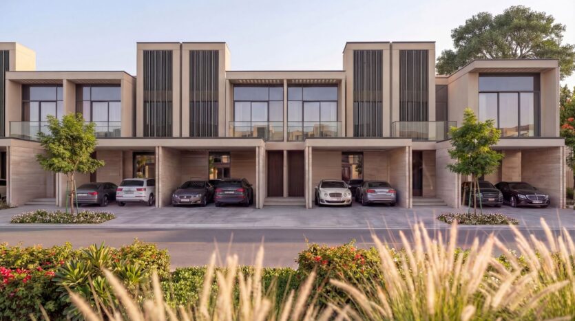 A modern row of two-story townhouses at The Terraces by Sobha, featuring large windows and covered driveways with parked cars. Lush landscaping, trees, grass, and vibrant flowers enhance the picturesque appeal of the foreground.