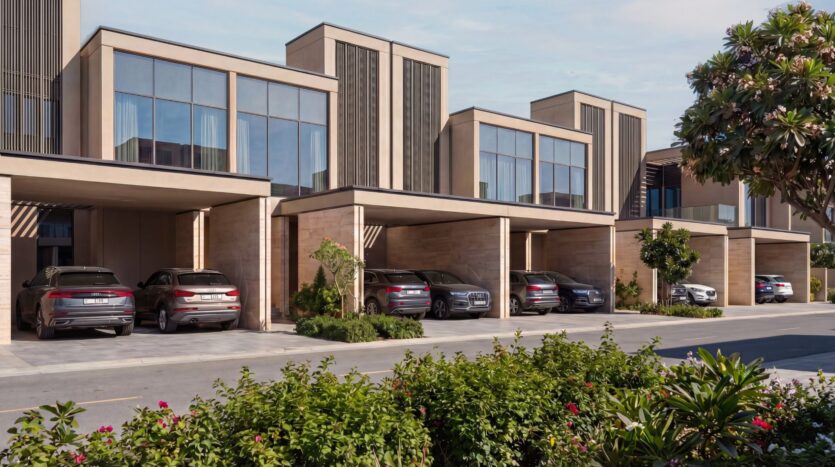 The modern townhouse complex The Terraces by Sobha features large floor-to-ceiling windows and carports. Several cars are parked in front of the house, while a landscaped green space with shrubs and trees stretches out in the foreground beneath a clear sky.