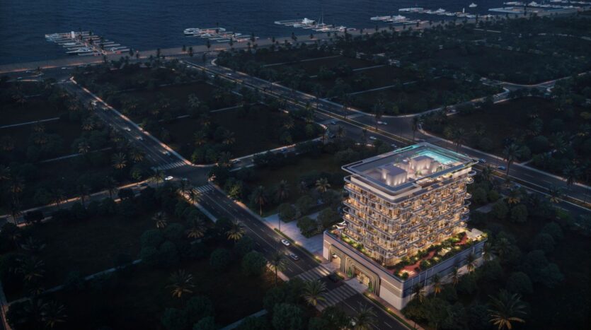 A nighttime aerial view of The Tides by Amis, a modern, multi-story building illuminated by lights and featuring a rooftop pool, surrounded by tree-lined streets and a picturesque waterfront with boats and piers in the background.