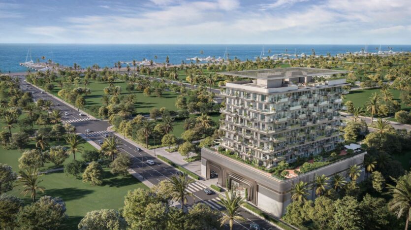 A modern multi-story building, The Tides by Amis, featuring numerous balconies, stands near a palm-lined road, overlooking a marina and the ocean. Green spaces and moored boats are visible in the background beneath a blue sky.