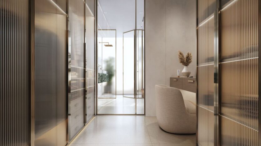 A modern, minimalist hallway with glass doors, beige walls, and built-in shelves features The Tides by Amis on the console table, alongside a light-colored armchair and a vase of dried flowers. The space is bathed in soft, natural light.