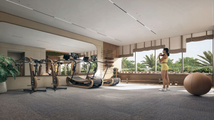 The modern fitness center at The Tides by Amis hotel is equipped with treadmills, large mirrors, potted plants, an exercise ball, and a woman in a yellow workout outfit drinking water near large windows overlooking palm trees.