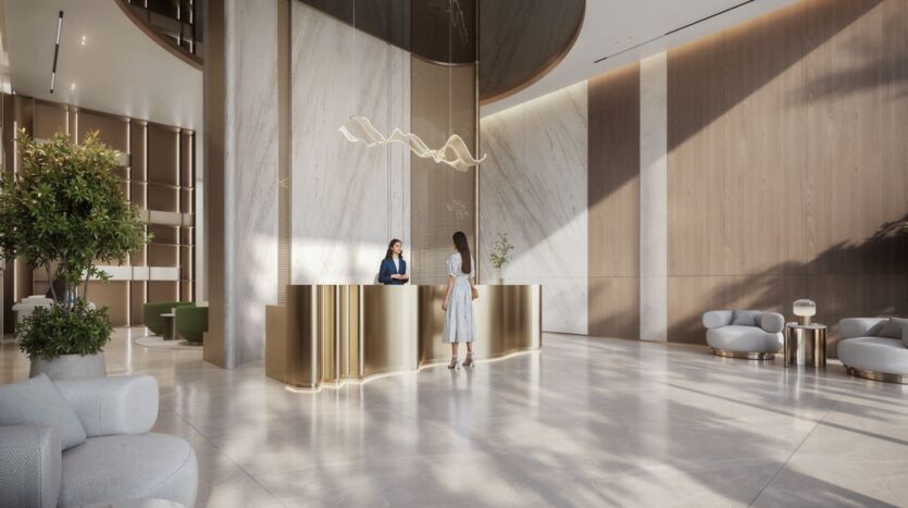 A modern hotel lobby with marble floors, a gold reception desk, and elegant lighting welcomes guests to The Tides by Amis. Two women are at the reception desk, surrounded by lush plants and stylish seating in a bright, spacious area.