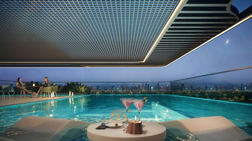 A luxurious rooftop pool at dusk at Tides by Amis, with a view of the city, two pink cocktails, and a dessert on a table in the water. Two people are sitting by the pool as a crescent moon shines in the clear evening sky.