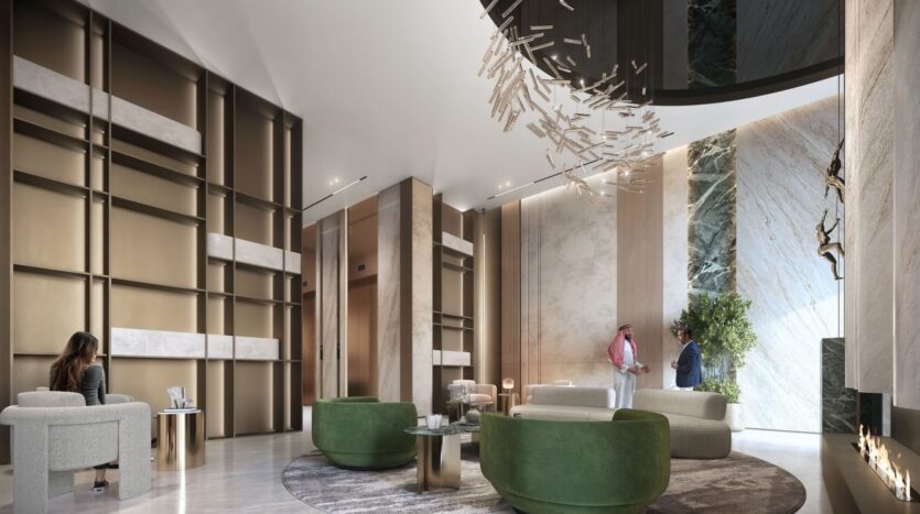 The lobby of a modern hotel features high ceilings, touches of gold and marble, and green circular chairs inspired by Amis’s *The Tides*. A unique abstract chandelier hangs above two people chatting near the elevator, while another person sits in a chair.