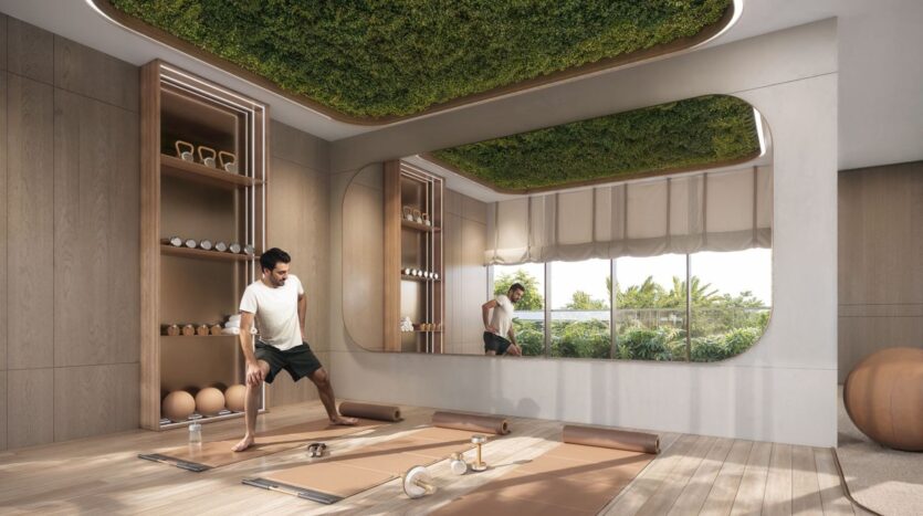 A man stretches in the bright, modern gym at The Tides by Amis, featuring wooden walls, a green ceiling, large mirrors, and neatly arranged dumbbells, mats, and kettlebells. Large windows offer a glimpse of the lush greenery outside.