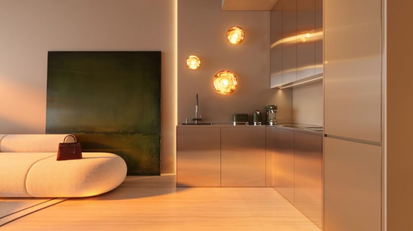 La cuisine moderne et minimaliste de la tour Wow est composée d'armoires métalliques, d'un canapé beige incurvé, d'un tableau vert foncé posé sur le sol et de trois appliques murales circulaires qui brillent chaleureusement. Un sac à main marron est posé sur le canapé.