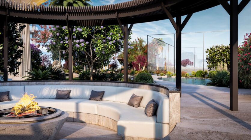 Le salon extérieur moderne de la Wow Tower est composé d'un canapé blanc incurvé avec des coussins gris sous une pergola, d'un foyer devant, d'une végétation luxuriante, de fleurs épanouies et d'une piscine avec une clôture en verre transparent à l'arrière-plan.