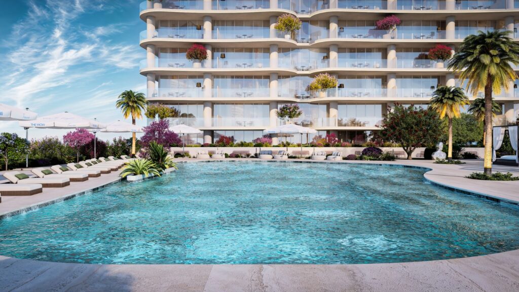 Une luxueuse piscine d'hôtel à l'eau bleue claire est nichée sous la tour Wow, entourée de chaises longues, de parasols, de palmiers et de fleurs colorées. Des balcons plantés de végétaux surplombent la piscine sous un ciel bleu parsemé de nuages.