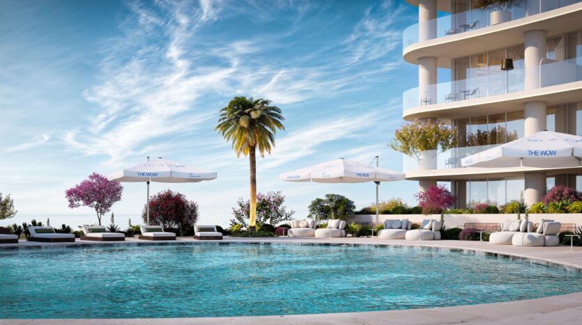La Wow Tower, un bâtiment moderne et incurvé doté de balcons, surplombe une piscine extérieure circulaire entourée de chaises longues, de parasols blancs, de palmiers et de plantes à fleurs éclatantes sous un ciel d'un bleu éclatant.