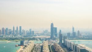 Una vista aérea envuelta en niebla de una ciudad moderna, con grandes rascacielos, grúas de construcción y edificios a orillas de unas aguas turquesas, pone de relieve el rápido crecimiento del sector inmobiliario de Dubái y su impacto en 2026 en esta dinámica zona urbana costera.