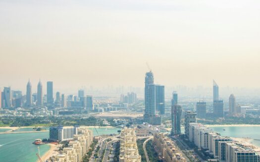 A misty aerial view of a modern city featuring towering skyscrapers, construction cranes, and buildings along a turquoise waterfront highlights the rapid growth of Dubai’s real estate sector and its impact on this dynamic coastal urban area by 2026.