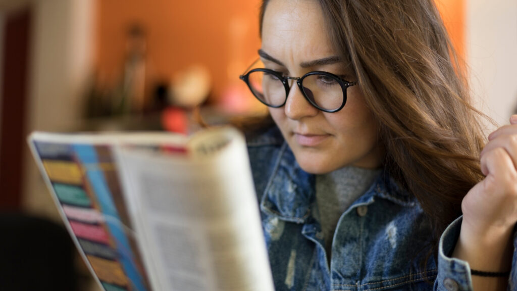 Eine Person mit Brille und Jeansjacke liest mit konzentriertem Gesichtsausdruck eine bunte Zeitschrift, die in Dubai Immo als „Challenges“ bezeichnet wird. Der Hintergrund ist in warmen Farbtönen leicht unscharf.