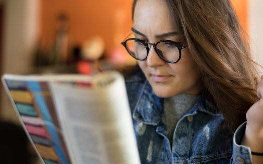 A person wearing glasses and a denim jacket is reading a colorful magazine, listed as "Challenges" in Dubai Immo, with a focused expression. The background is slightly blurred in warm tones.
