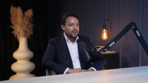 Un hombre vestido con traje está sentado a una mesa y habla por un micrófono durante el programa Podcast SUCCESS. En el fondo se ve un jarrón decorativo con plantas secas y una lámpara encendida, lo que crea un ambiente moderno y profesional.