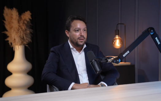Ein Mann im Anzug sitzt an einem Tisch und spricht während der Podcast-Sendung „SUCCESS“ in ein Mikrofon. Im Hintergrund stehen eine dekorative Vase mit Trockenblumen und eine eingeschaltete Lampe, die einen modernen und professionellen Rahmen schaffen.