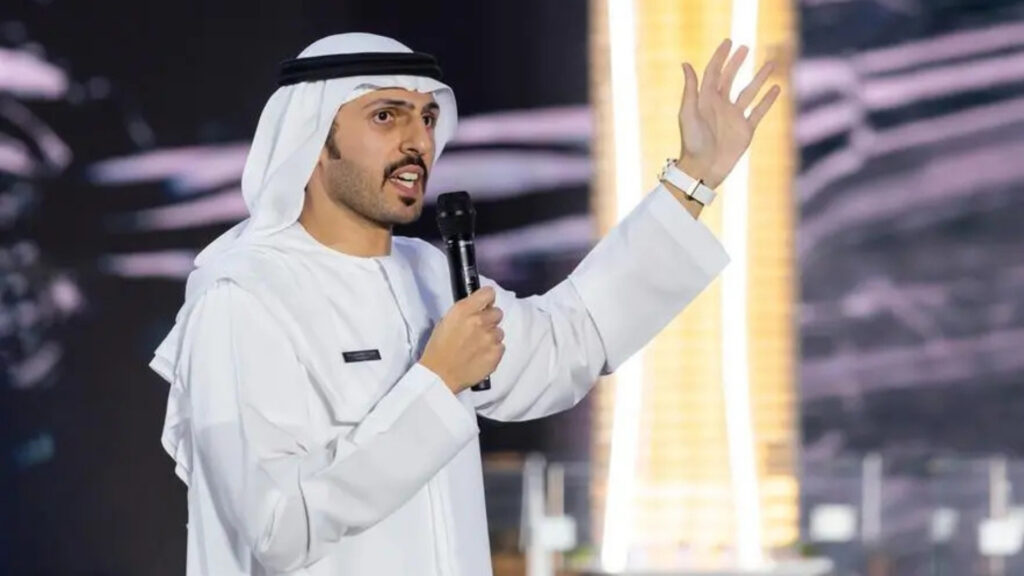Ein Mann in traditioneller Kleidung aus dem Nahen Osten spricht mit erhobener Hand vor einem unscharfen, beleuchteten Hintergrund aus Dubai in ein Mikrofon und spricht möglicherweise über die Finanzierung von Bauträgern oder andere finanzbezogene Themen.