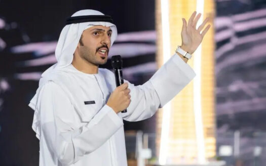 Ein Mann in traditioneller Kleidung aus dem Nahen Osten spricht mit erhobener Hand vor einem unscharfen, beleuchteten Hintergrund aus Dubai in ein Mikrofon und spricht möglicherweise über die Finanzierung von Bauträgern oder andere finanzbezogene Themen.