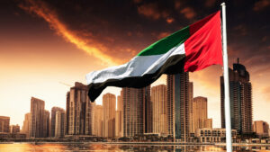En primer plano ondea la bandera de los Emiratos Árabes Unidos, con modernos rascacielos y una espectacular puesta de sol sobre un paisaje urbano: una imagen muy popular entre los inversores que buscan un visado de residencia en Dubái y se informan sobre la cantidad mínima requerida.