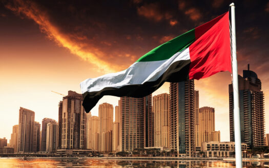 The flag of the United Arab Emirates flies in the foreground, with modern skyscrapers and a spectacular sunset over the cityscape—a popular image among investors seeking a Dubai residency visa and researching the minimum investment requirement.