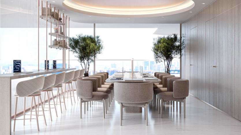 Salle à manger moderne avec des chaises beiges autour d'une longue table, de grands tabourets de bar sur un comptoir à gauche, et deux arbres en pot. Des fenêtres allant du sol au plafond offrent une vue lumineuse sur le paysage urbain. L'élégance minimaliste inspirée de Cybele by Wadan remplit l'espace naturellement éclairé.