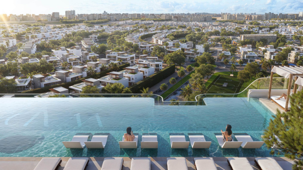 La piscine à débordement sur le toit du Cybele by Wadan, où des personnes se prélassent, surplombe un paysage urbain tentaculaire de villas blanches modernes et de verdure luxuriante sous un ciel bleu.
