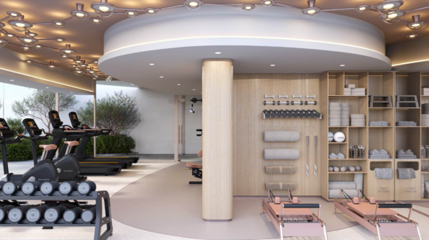Une salle de sport moderne avec des tapis de course Cybele by Wadan, des poids libres, des réformateurs de pilates et des étagères contenant des serviettes, des tapis de yoga et des équipements de fitness. L'espace est agrémenté de grandes fenêtres, de plantes et d'un éclairage élégant au plafond.