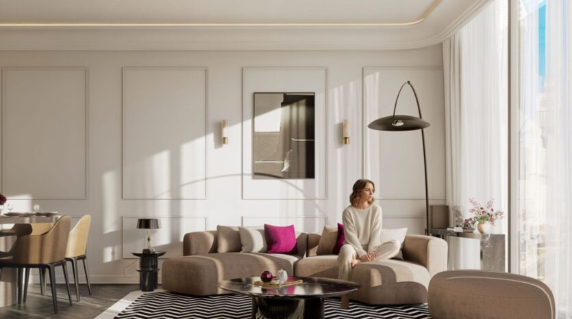 Une femme est assise sur un canapé beige moderne dans le salon lumineux et stylé de la résidence Fauchon, doté de grandes fenêtres, de murs neutres, d'un lampadaire noir et de coussins d'appoint roses. La lumière du soleil entre à flots et illumine l'espace.