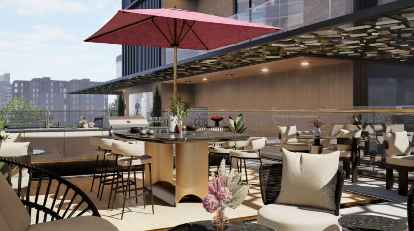 Un patio extérieur moderne de la résidence Fauchon avec des chaises et des tables beiges, un grand barbecue rond et un parasol rouge. Les bâtiments de la ville sont visibles à l'arrière-plan et des plantes en pot ajoutent de la verdure à ce cadre élégant.