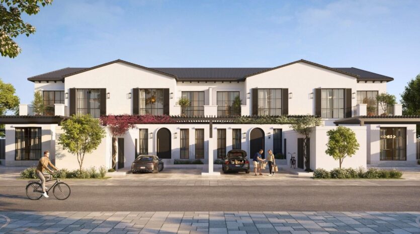 Complexe moderne de maisons de ville blanches de deux étages avec de grandes fenêtres, des balcons et des arches à Hudayriyat Golf Estates. Deux voitures sont garées devant, des gens se tiennent près de l'entrée et une personne fait du vélo sur les trottoirs bordés d'arbres.