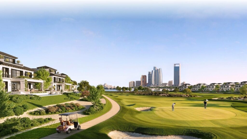Un parcours de golf pittoresque à Hudayriyat Golf Estates montre deux personnes sur un green, entourées de maisons modernes et de verdure luxuriante. Une voiturette de golf suit un chemin tandis qu'une ligne d'horizon de la ville avec de grands bâtiments s'élève à l'arrière-plan sous un ciel clair.