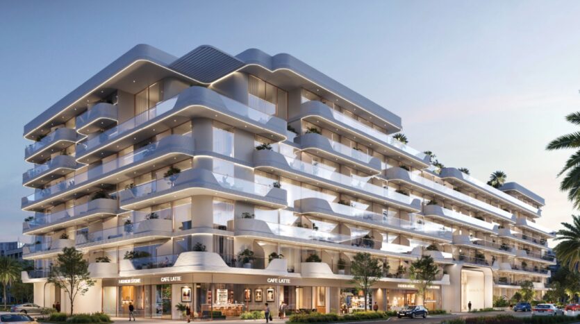 Un immeuble moderne de plusieurs étages - Oxford Cove by Iman - est doté de balcons blancs et épurés, de garde-corps en verre et de palmiers sur les terrasses. Des boutiques et des cafés bordent le rez-de-chaussée, avec des voitures garées et des gens qui se promènent à proximité au coucher du soleil.
