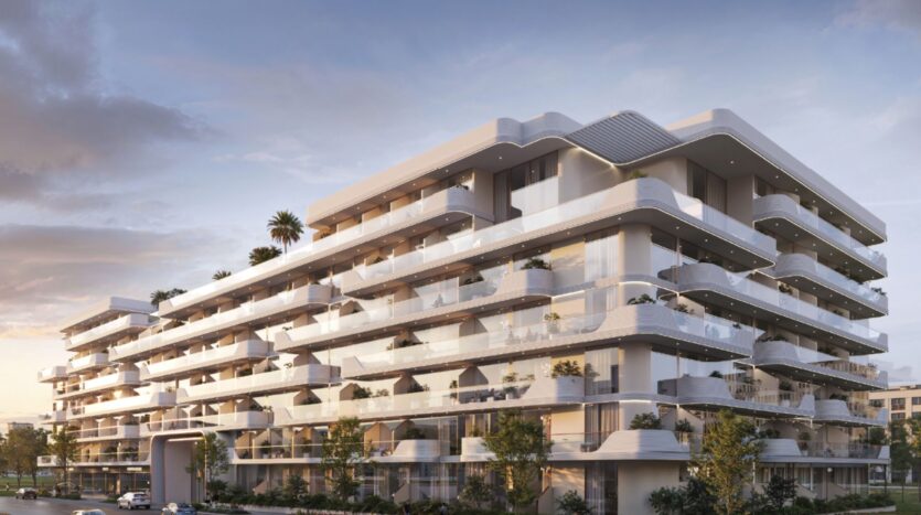 Oxford Cove by Iman est un immeuble d'appartements moderne de plusieurs étages, doté de grands balcons, de garde-corps en verre et d'éléments architecturaux courbes, entouré de verdure et de palmiers sous un ciel nuageux au coucher du soleil.