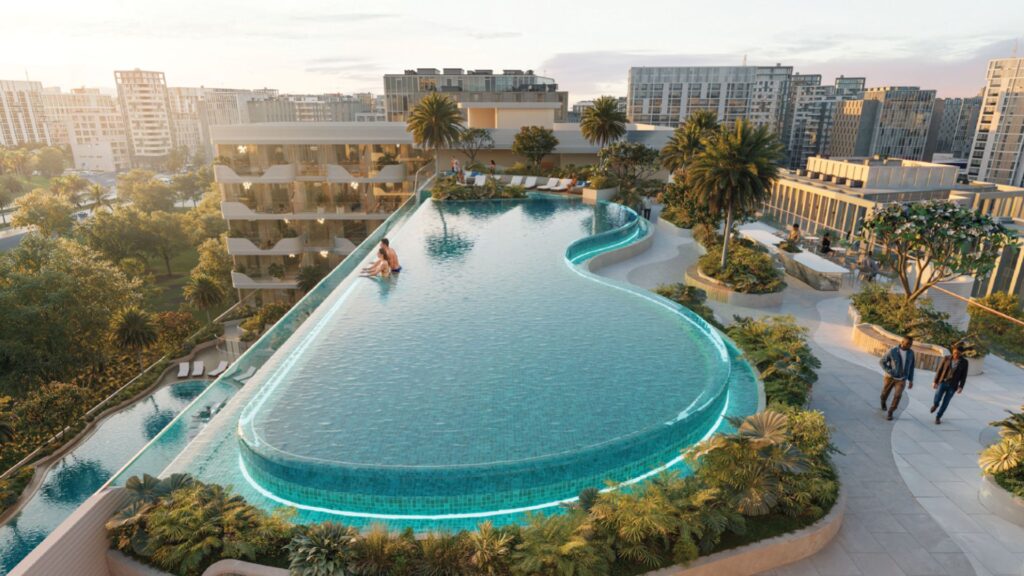 La piscine à débordement d'Oxford Cove by Iman, dont les bords sont d'un bleu éclatant, surplombe un paysage urbain composé d'immeubles modernes et de palmiers, tandis que les gens se détendent au bord de l'eau au coucher du soleil.