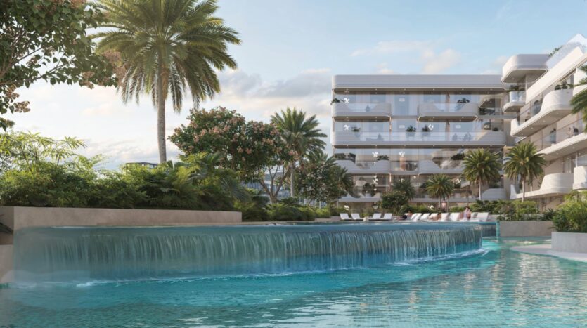 L'immeuble d'appartements de luxe moderne Oxford Cove by Iman est doté de balcons en verre donnant sur une piscine sereine avec une cascade, entourée d'une végétation luxuriante, de palmiers et de chaises longues sous un ciel bleu.