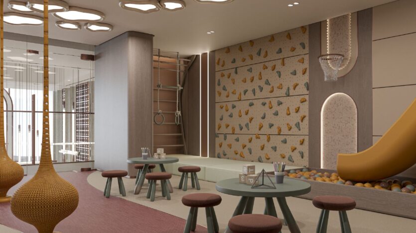 Une salle de jeux intérieure moderne à Oxford Cove par Iman comprend de petites tables rondes et des tabourets, un toboggan se terminant par une fosse à balles, un mur d'escalade, des anneaux de gymnastique et un panier de basket-ball. Les tons chauds et les plafonniers décoratifs créent une atmosphère chaleureuse.