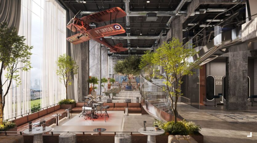L'intérieur moderne et industriel du Raw District by Imtiaz présente de grandes fenêtres, de grands arbres et des sièges de salon. Un biplan rouge d'époque est suspendu au-dessus d'une scène avec une batterie, encadrée par des murs en verre et un décor contemporain.